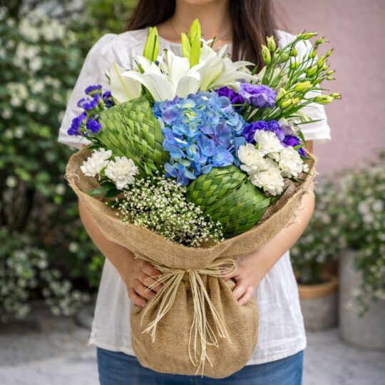 Blue hydrangea bouquet with white lilies, carnations and woven leaf detailing wrapped in jute by FreshKnots Bangalore with same day delivery.