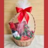 Christmas gift basket with gourmet snacks, a holiday beverage, chocolates, and festive décor, wrapped with white net and a red bow.