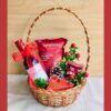 Christmas gift basket with gourmet snacks, a holiday beverage, chocolates, and festive décor, wrapped with white net and a red bow.