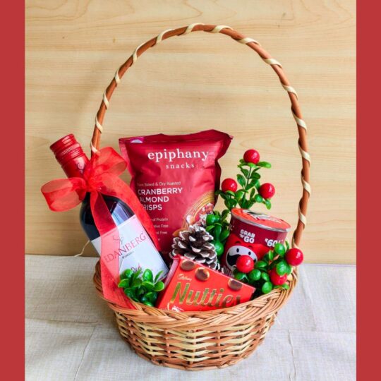 Christmas gift basket with gourmet snacks, a holiday beverage, chocolates, and festive décor, wrapped with white net and a red bow.