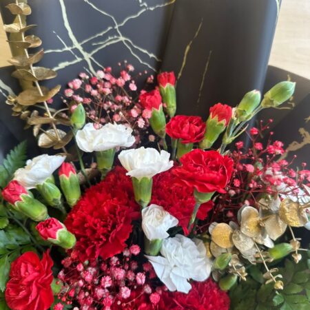 Christmas-themed bouquet with red carnations, white flowers, greens, and gold accents in black wrapping.