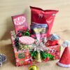 Christmas treat hamper with chocolates, popcorn, nuts, beverage can and festive décor placed in a red Christmas box.