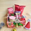 Christmas treat hamper with chocolates, popcorn, nuts, beverage can and festive décor placed in a red Christmas box.