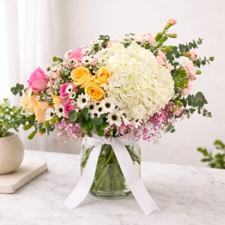 A pastel floral arrangement in a glass vase featuring white hydrangeas, yellow and pink roses, white daisies, and pink fillers, tied with a white ribbon - ideal for same day flower delivery in Bangalore.