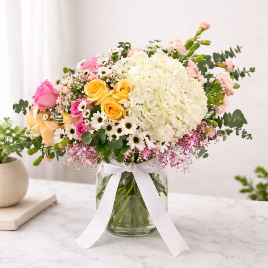 A pastel floral arrangement in a glass vase featuring white hydrangeas, yellow and pink roses, white daisies, and pink fillers, tied with a white ribbon - ideal for same day flower delivery in Bangalore.