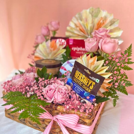 Gift hamper with chocolates, pink roses, baby’s breath, and a candle arranged in a woven basket with a pink ribbon