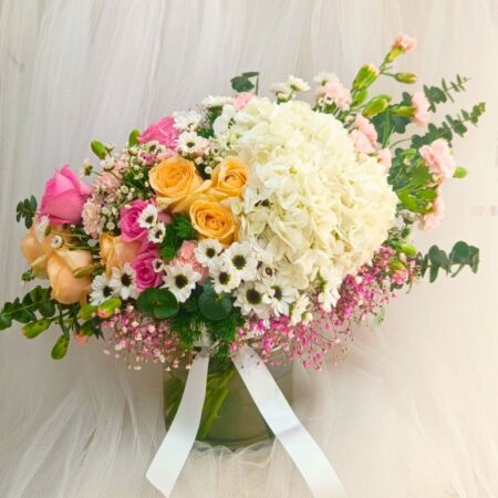 A pastel floral arrangement in a glass vase featuring white hydrangeas, yellow and pink roses, white daisies, and pink fillers, tied with a white ribbon — ideal for same day flower delivery in Bangalore.