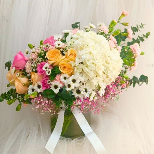 A pastel floral arrangement in a glass vase featuring white hydrangeas, yellow and pink roses, white daisies, and pink fillers, tied with a white ribbon — ideal for same day flower delivery in Bangalore.