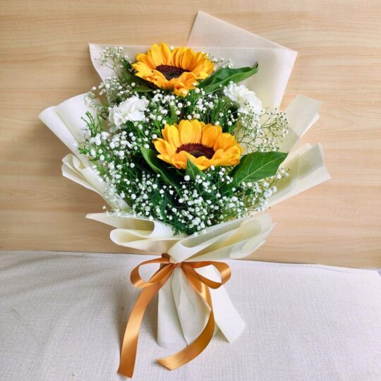 A cheerful sunflower bouquet paired with white carnations and baby’s breath, elegantly wrapped in cream paper with a golden ribbon — available for same day flower delivery in Bangalore.