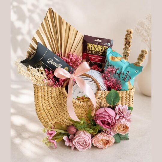 Luxury woven gift hamper with chocolates, snacks, and pink artificial flowers from FreshKnots Bangalore.