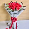 A bouquet of fresh red roses wrapped in white and black paper with baby’s breath and a red bow, placed with scattered rose petals around it.
