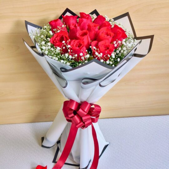 A bouquet of fresh red roses wrapped in white and black paper with baby’s breath and a red bow, placed with scattered rose petals around it.