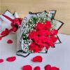A bouquet of fresh red roses wrapped in white and black paper with baby’s breath and a red bow, placed with scattered rose petals around it.