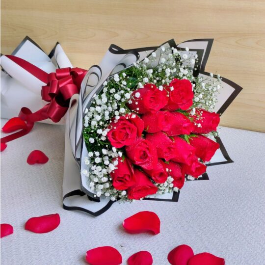 A bouquet of fresh red roses wrapped in white and black paper with baby’s breath and a red bow, placed with scattered rose petals around it.