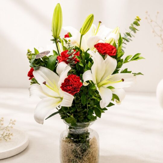A glass vase arrangement of white lilies and red carnations with lush green foliage - ideal for same day flower delivery in Bangalore.