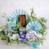 Blue themed gourmet gift hamper with chocolates, cookies, snacks and artificial flowers arranged in a wooden tray.