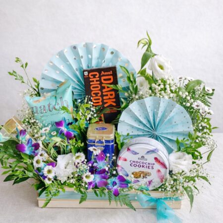 Blue themed gourmet gift hamper with chocolates, cookies, snacks and artificial flowers arranged in a wooden tray.