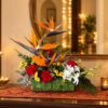 Tall bird of paradise stems with red carnations, yellow chrysanthemums and white orchids in a handwoven base, angled view