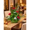 Red rose and carnation centrepiece in a woven leaf basket on a dining table runner, angled view