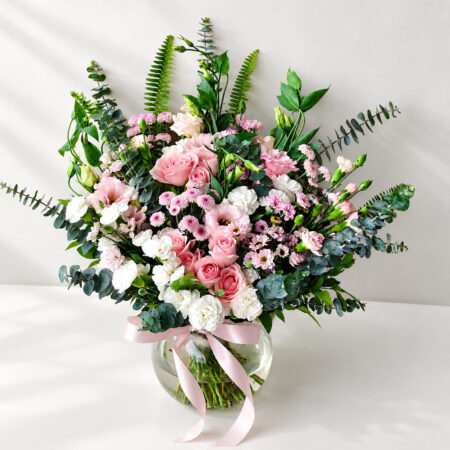 Pastel pink and white flower arrangement with roses and eucalyptus in a round glass vase with pink ribbon, angled view
