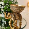 Close-up of an antique gold metal elephant candle holder with an engraved bowl, beside white and green chrysanthemums