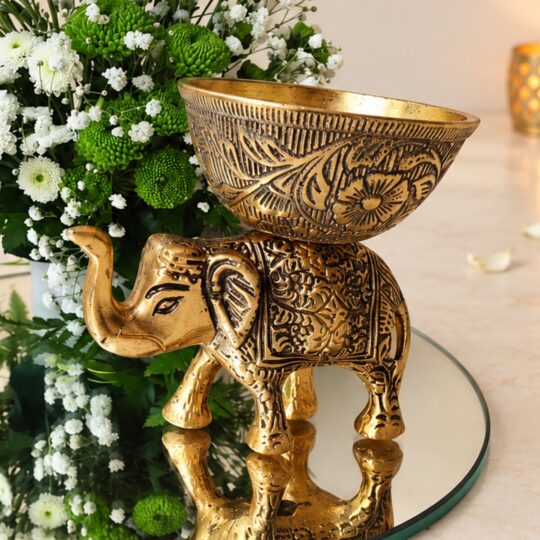 Close-up of an antique gold metal elephant candle holder with an engraved bowl, beside white and green chrysanthemums